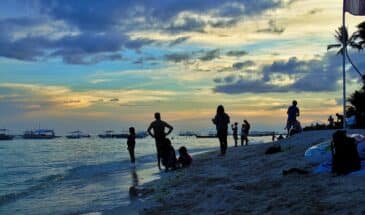 Plage Philippines : sous le soleil couchant, des touristes sont sur le sable, il y a des bateaux sur la mer.