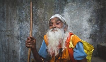 Vieil homme Népal : il y a un vieil homme assis contre une pierre qui tient une canne en bois et porte un sac jaune.