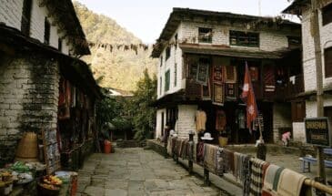 Village Népal : on voit des immeubles en pierre, un stand de nourriture, des tapis et des foulards sont étendus.