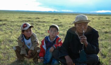 Culture Mongolie : un homme sénior portant un chapeau avec deux enfants assis à coté de lui dans de l'herbe.