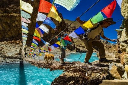 Trek Markha : des randonneurs avec un chien traversant un pont en bois, des cordes de couleurs et un lac.